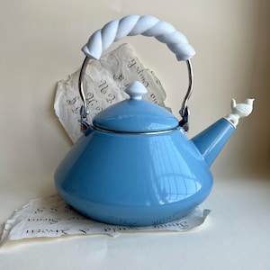 May include: A light blue enamel teapot with a white twisted handle and a small white bird figurine on the spout. The teapot has a silver metal frame and a white knob on the lid. The teapot is sitting on a piece of paper with handwritten text.