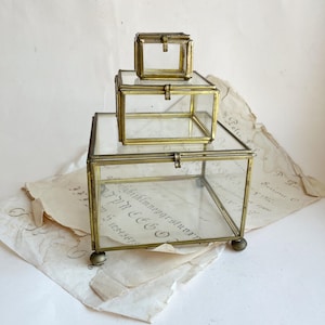Könnte beinhalten: Drei Vintage-Glas- und Messingboxen unterschiedlicher Größe sind übereinander gestapelt. Die Boxen haben eine rechteckige Form mit Scharnierdeckeln und kleinen Kugelfüßen. Das Glas ermöglicht einen Blick auf den Inhalt, und die Messingrahmen verleihen Eleganz.