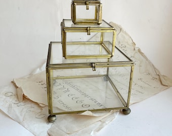 Könnte beinhalten: Drei Vintage-Glas- und Messingboxen unterschiedlicher Größe sind übereinander gestapelt. Die Boxen haben eine rechteckige Form mit Scharnierdeckeln und kleinen Kugelfüßen. Das Glas ermöglicht einen Blick auf den Inhalt, und die Messingrahmen verleihen Eleganz.