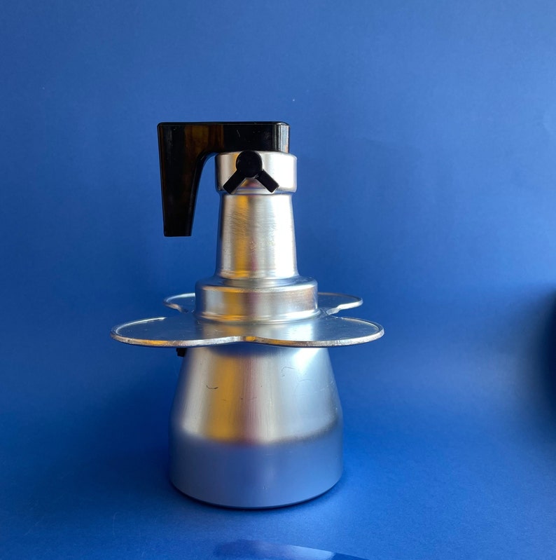 Op de afbeelding: Een zilveren espressoapparaat voor op het fornuis met een zwarte kunststof handgreep. Het apparaat heeft een drietandvormige basis.