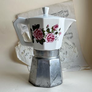 Può includere: Una caffettiera moka con la parte superiore in ceramica bianca decorata con motivi di rose rosa e un coperchio abbinato, fissata su una base ottagonale in alluminio metallico. Il design presenta un manico e un beccuccio per versare.