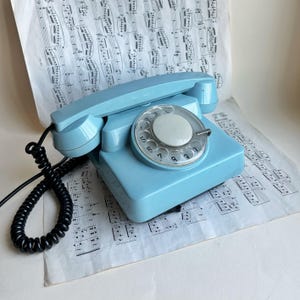 May include: A light blue rotary telephone with a circular dial featuring numbers and a white center. The handset rests on the base, connected by a black coiled cord. The device has a smooth, glossy finish and a classic rectangular base design.