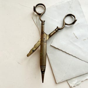 May include: Vintage brass scissors with looped handles and a pointed tip. The scissors have a worn, antique appearance. The blades are slightly open, suggesting they are ready for use. The scissors rest on a white surface with paper. The brass has a tarnished finish.