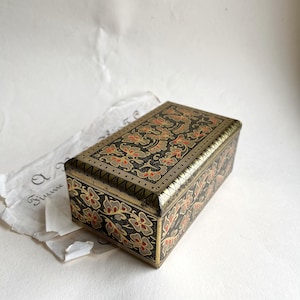 Op de afbeelding: Een rechthoekige zwarte doos met een bloemmotief in rood en goud. De doos heeft een gouden rand en ligt op een wit oppervlak. Een stuk papier met cursief schrift ligt naast de doos.