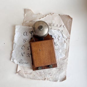 Puede incluir: Un timbre de pared con una cúpula de metal plateado pulido fijada a una base rectangular de madera. El soporte de madera tiene un acabado marrón oscuro e incluye dos pequeños detalles metálicos circulares en la parte inferior y un brazo percutor.