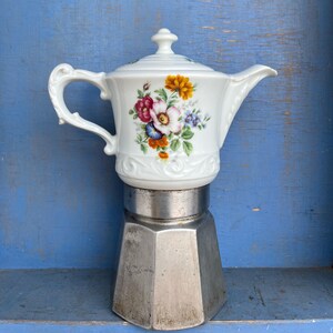 Puede incluir: Una tetera de porcelana blanca con un ramo de flores rojas, amarillas y azules, rematada con una tapa. La tetera está unida a una base de aluminio metálico de forma octogonal, creando una cafetera híbrida con un asa curva.
