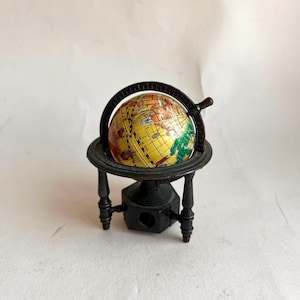Pode incluir: Um pequeno apontador de lápis em forma de globo. O globo é amarelo com continentes vermelhos, verdes e castanhos. Está num suporte de metal preto com uma pega e uma abertura para o apontador. O suporte tem quatro pernas.
