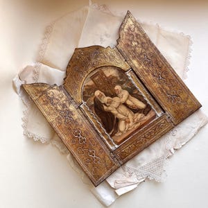 May include: An antique, gold-toned triptych with a religious painting at the center. The painting depicts figures in a somber scene. The triptych is open, revealing ornate, embossed designs on the side panels. The piece rests on a stack of vintage lace and paper.