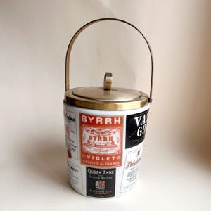 May include: Vintage ice bucket with a white ceramic body and a metal lid and handle. The bucket is adorned with various liquor labels, including "BYRRH" and "V.A. 69". The handle arches over the top, and the lid has a small ring for lifting.