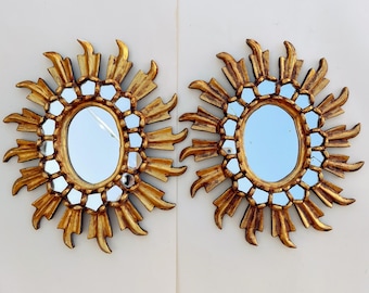 May include: A pair of gold sunburst mirrors with oval-shaped glass mirrors in the center. The mirrors are surrounded by a decorative design of gold rays.