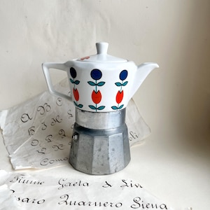 May include: A white ceramic coffee pot with a floral pattern in red, blue, and green, atop a silver-coloured metal base. The coffee maker sits on paper with handwritten text. A classic Italian design.