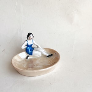 May include: A small, oval-shaped dish with a seated figurine. The figurine is a woman in a blue swimsuit, with black shoes and hair. The dish is a light beige color.