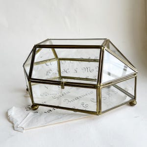 May include: A clear glass and brass geometric box with a hinged lid and small brass feet. The box has a decorative design and is likely used for storing small items. A piece of paper with script is beneath the box.