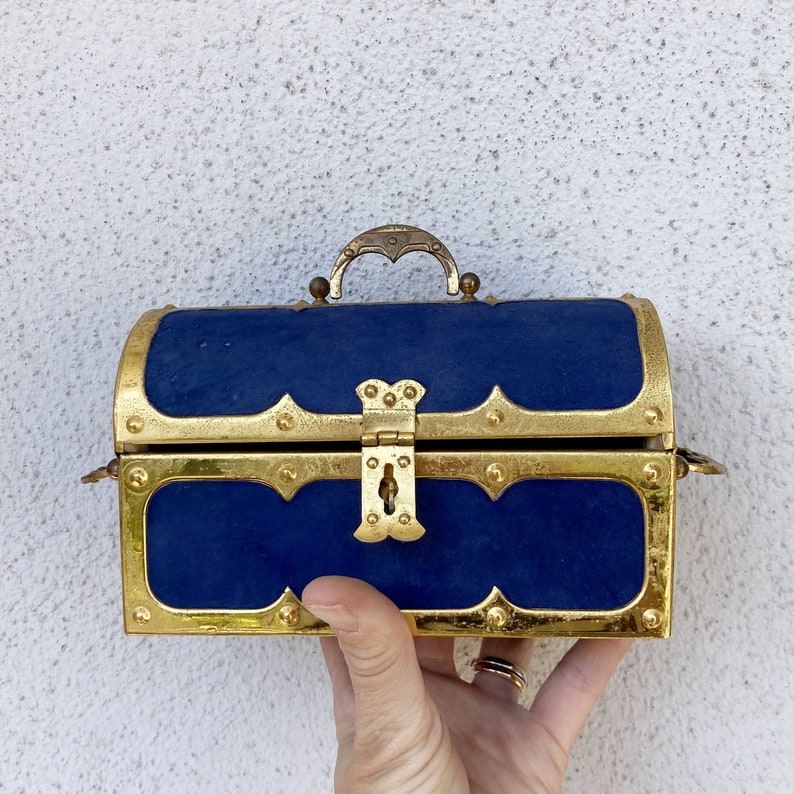 May include: A small, gold-colored treasure chest with a blue velvet lining. The chest has a hinged lid with a decorative lock and a handle.