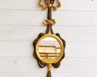 May include: A gold and black ornate wall mirror with a tassel hanging from the bottom. The mirror has a round shape with a decorative frame and a bow at the top.