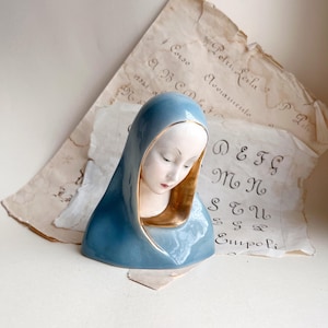 May include: A ceramic bust of a woman with a serene expression, wearing a blue veil with gold trim. The face is pale with delicate features and closed eyes. The bust rests on a surface with aged paper and handwritten script in the background.