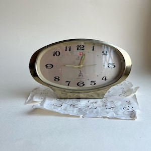 May include: Oval-shaped, vintage alarm clock with a gold-colored frame and a light pink face. The clock features black numerals, gold hands, and the text "FIVE" and "MADE IN CHINA". The clock sits on a piece of paper.