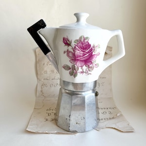 Könnte beinhalten: Eine Vintage-Espressokanne mit einem weißen Keramikaufsatz mit einem rosafarbenen Rosendesign. Der Sockel ist aus silberfarbenem Metall und der Griff ist schwarz. Die Kaffeemaschine steht auf einem Stück gealtertem Papier.