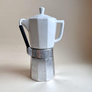 Pode incluir: Uma cafeteira italiana com uma parte superior de cerâmica branca, incluindo tampa e pega a condizer. A base inferior é feita de metal prateado escovado. Uma pega cilíndrica preta está fixada na lateral da unidade.