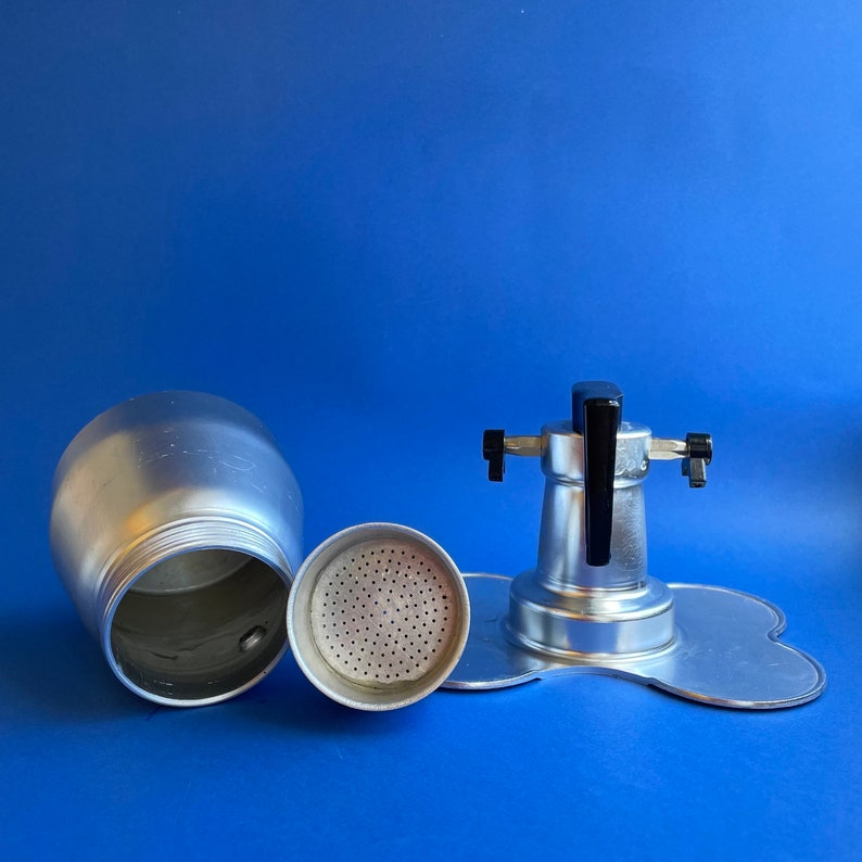 Op de afbeelding: Een zilveren aluminium koffiezetapparaat met een zwarte handgreep en een verwijderbaar filter. De koffiezetapparaat is gedemonteerd, waardoor de afzonderlijke onderdelen zichtbaar zijn.