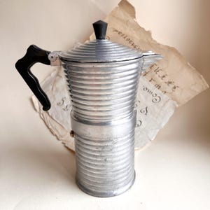 Puede incluir: Una cafetera italiana plateada vintage con asa y tapa negras. La cafetera tiene un diseño acanalado y está colocada sobre un fondo de papel envejecido con texto manuscrito. Un electrodoméstico de cocina clásico.