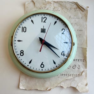 Puede incluir: Un reloj de pared redondo con un marco verde pálido y una esfera blanca. El reloj presenta números arábigos negros a intervalos regulares, marcadores de hora negros en forma de diamante, manecillas negras y un segundero rojo fino. El nombre de la marca Melux está impreso en negro sobre el centro.