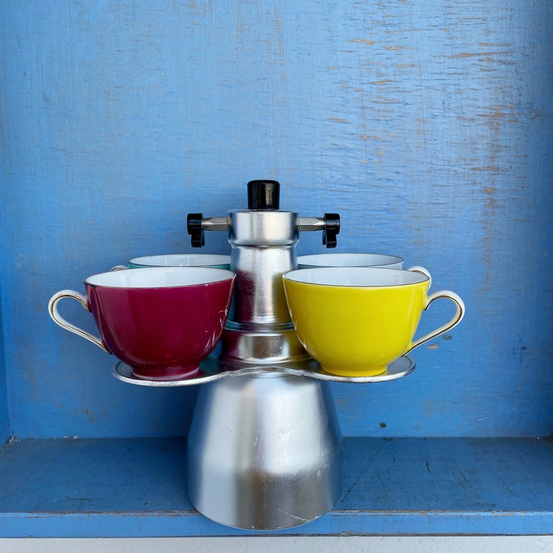 Op de afbeelding: Een zilveren koffiezetapparaat met vier kopjes, waarvan er twee rood en geel zijn. De kopjes staan op een zilveren dienblad.