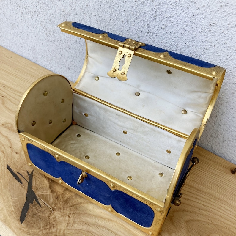 May include: A small, blue and gold treasure chest with a hinged lid and a brass lock. The chest is lined with white fabric and has a decorative brass handle.