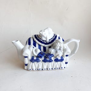 Peut inclure: Théière en céramique blanche avec des rayures et des accents bleus. La théière présente un motif d'une personne assise sur un canapé avec un chat au-dessus. Le canapé a des livres bleus et un chapeau. La théière a un bec et une anse.