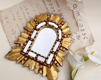 Puede incluir: Un pequeño espejo dorado y ornamentado con un marco en forma de sol. El espejo tiene forma rectangular con la parte superior arqueada y está rodeado por un borde decorativo y texturizado. Se encuentra sobre un papel antiguo con texto manuscrito y acompañado de lirios cala blancos.