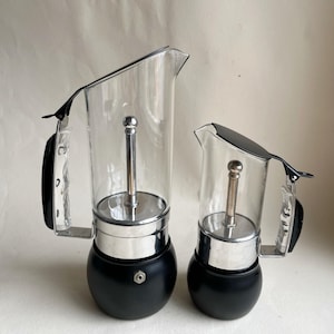 Pode incluir: Duas cafeteiras de espresso de vidro. As cafeteiras têm uma base preta, uma seção intermediária prateada e um topo de vidro transparente com uma alça preta. As cafeteiras de espresso são de tamanhos diferentes.