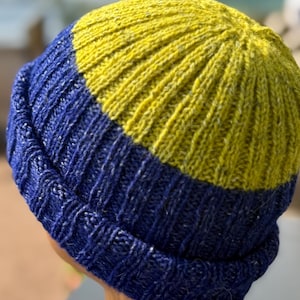 May include: A knitted beanie hat in a two-tone color scheme. The lower portion is a deep blue, while the upper section is a vibrant yellow-green. The hat is made of a textured yarn, and the ribbed design is visible.