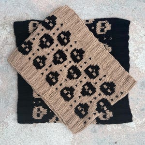 Könnte beinhalten: Zwei gestrickte Schals, einer in Beige und einer in Schwarz, werden gezeigt. Der beige Schal mit einem sich wiederholenden schwarzen Muster ist über dem schwarzen Schal angeordnet. Das Muster scheint ein stilisiertes Design zu sein.
