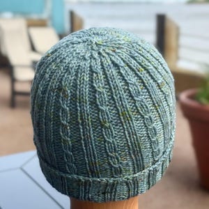 Puede incluir: Un gorro tejido a mano en tonos azules y verdes con un diseño de punto acanalado y trenzado. El gorro tiene un borde doblado y una apariencia texturizada. El hilo tiene sutiles motas amarillas y verdes.