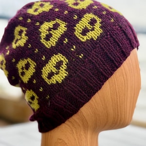Può includere: Un berretto lavorato a maglia di colore viola scuro, adornato con un motivo ripetuto di teschi gialli. Il cappello è esposto su una forma di testa in legno, che ne mette in risalto la forma e la consistenza. Il motivo a teschio aggiunge un tocco unico a questo accessorio invernale.