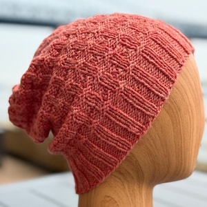 Puede incluir: Un gorro tejido de color coral con un patrón de diamantes texturizado. El gorro se muestra en una cabeza de madera, mostrando su ajuste holgado y el borde acanalado. El gorro está hecho de hilo suave.