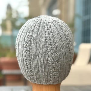 May include: A gray knitted beanie hat with a ribbed design and textured accents. The hat is made of a soft, possibly wool, material. The hat is shown on a wooden form.