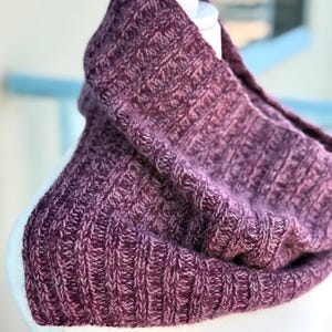 Puede incluir: Un cuello tejido en tonos morados y rosas, exhibido en una cabeza de maniquí blanca. El cuello presenta una textura acanalada y parece suave y cálido, perfecto para el clima frío. El primer plano resalta el intrincado patrón de tejido.