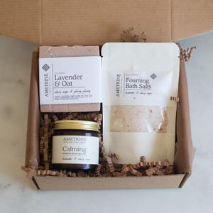 May include: A brown cardboard gift box containing self-care products. The box includes a lavender and oat soap bar, foaming bath salts, and a jar of whipped body butter. All items are labelled with the brand "Ametrine" and the word "Calming".