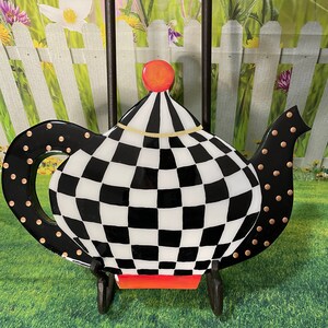 May include: A black and white checkered teapot-shaped ceramic dish with a red and gold top. The dish has gold polka dots around the rim.