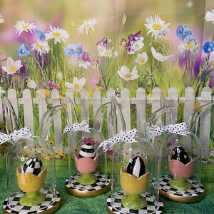 May include: Five glass cloche Easter egg decorations with bunny ears. Each cloche features a black and white egg in a yellow, pink, or green tulip base. The bases are on a black and white checkered surface.