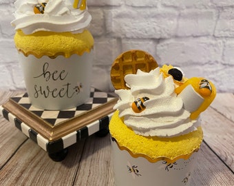 May include: Two yellow and white bee-themed cupcakes with waffle toppers. The cupcake on the left has a white wrapper with the text "bee sweet" and the cupcake on the right has a blue wrapper with a bee pattern.