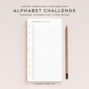 Könnte beinhalten: Ein weißer Planereinsatz mit dem Text "ALPHABET CHALLENGE" und linierten Feldern für jeden Buchstaben des Alphabets. Ein Bleistift aus Holz mit schwarzem Radiergummi liegt neben dem Planer. Der Planer ist für den persönlichen Gebrauch bestimmt.
