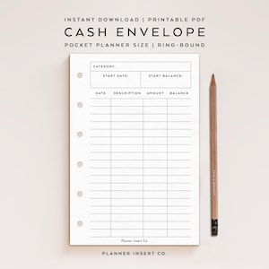 May include: A white cash envelope planner insert with black grid lines and text. The text includes "CASH ENVELOPE", "POCKET PLANNER SIZE", and fields for date, description, amount, and balance. A wooden pencil rests on the right side.