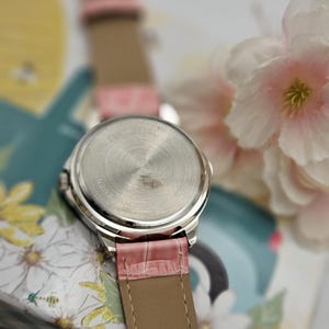 May include: A silver-toned wristwatch with a pink leather band. The watch face is not visible, but the stainless steel back is in focus. The band is a light pink with a textured, crocodile-like pattern. The watch is set against a floral background.