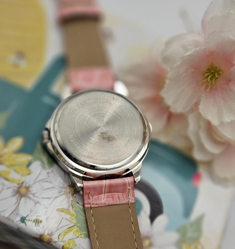 May include: A silver-toned wristwatch with a pink leather band. The watch face is not visible, but the stainless steel back is in focus. The band is a light pink with a textured, crocodile-like pattern. The watch is set against a floral background.