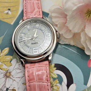 May include: A silver quartz watch with a light pink faux-leather band. The watch face is white with silver hands and numerals at 12, 3, and 6 o'clock. The band has a crocodile skin texture. The watch is on a floral background.