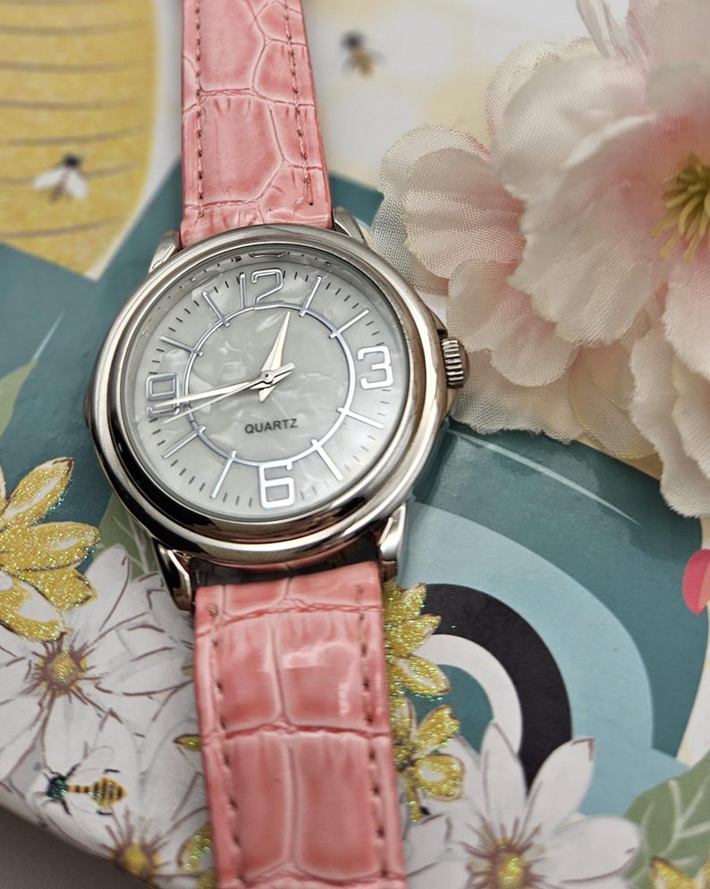 May include: A silver quartz watch with a light pink faux-leather band. The watch face is white with silver hands and numerals at 12, 3, and 6 o'clock. The band has a crocodile skin texture. The watch is on a floral background.