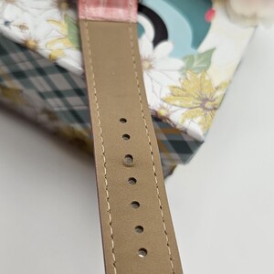 May include: Close-up of a wristwatch with a pink leather band and a silver-colored watch face. The band is stitched with a light-colored thread. The watch is resting on a floral patterned box.
