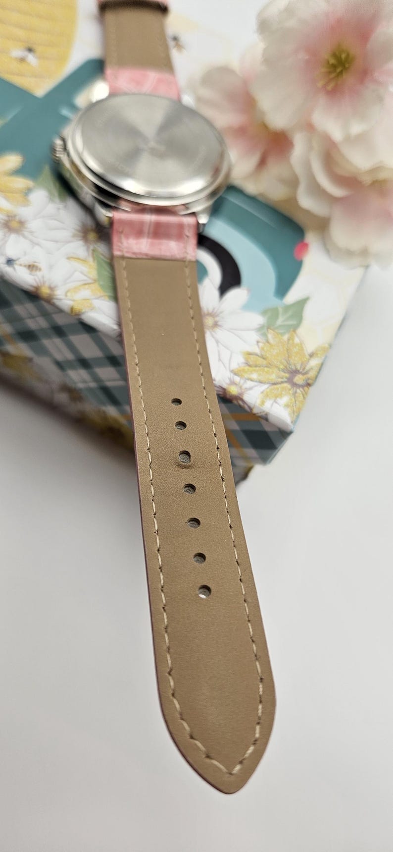 May include: Close-up of a wristwatch with a pink leather band and a silver-colored watch face. The band is stitched with a light-colored thread. The watch is resting on a floral patterned box.